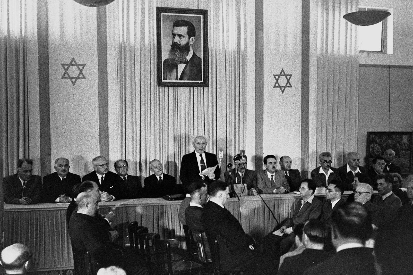 David Ben Gurion vyhlašuje nezávislost Izraele. Na obrazu nad ním je Theodor Herzl, „duchovní otec sionismu“.